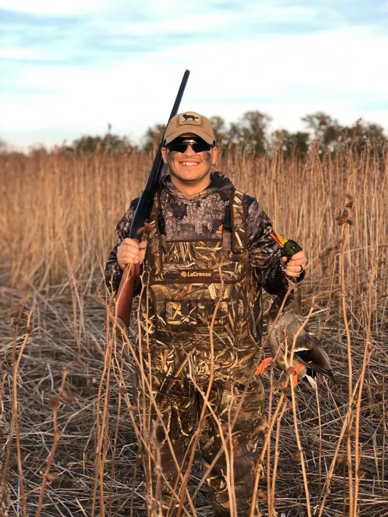 A smiling hunter in full camouflage, holding a shotgun and two harvested ducks, stands in a field of tall, dry reeds.