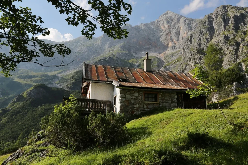 A rustic stone and wood cabin with a red metal roof nestled on a green, grassy hillside, surrounded by trees and majestic, rocky mountains under a partly cloudy sky, conveying an outdoor, remote lifestyle.