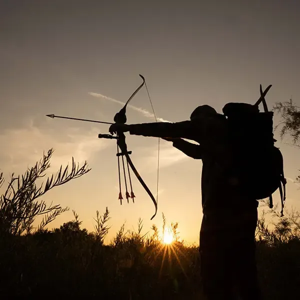 A silhouette of a bowhunter aiming a recurve bow with an arrow nocked, wearing a backpack, against a vibrant sunset sky in a natural, grassy landscape.