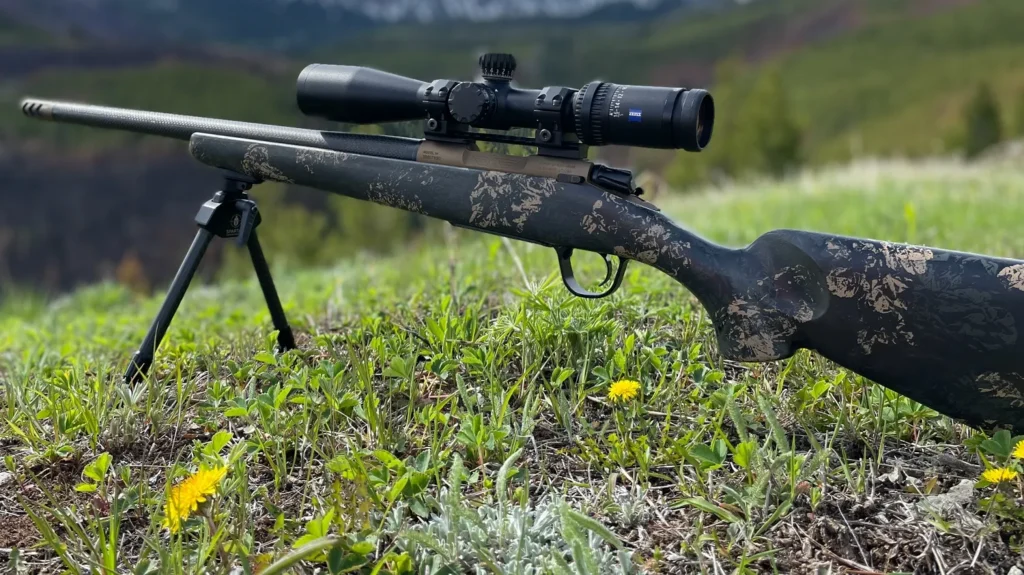 A Christensen Arms Ridgeline FFT rifle with a camouflaged carbon fiber stock and a large scope, propped up on a bipod in a grassy, outdoor setting with a mountain backdrop.