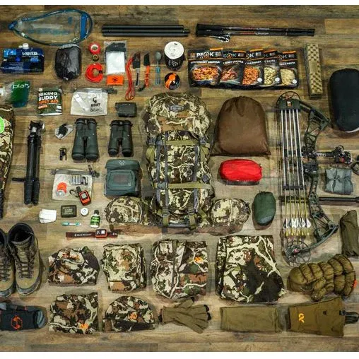 An overhead flat lay of a complete hunting gear setup, including camouflage clothing, a compound bow, binoculars, survival tools, and dehydrated food, all neatly arranged on a wooden surface.