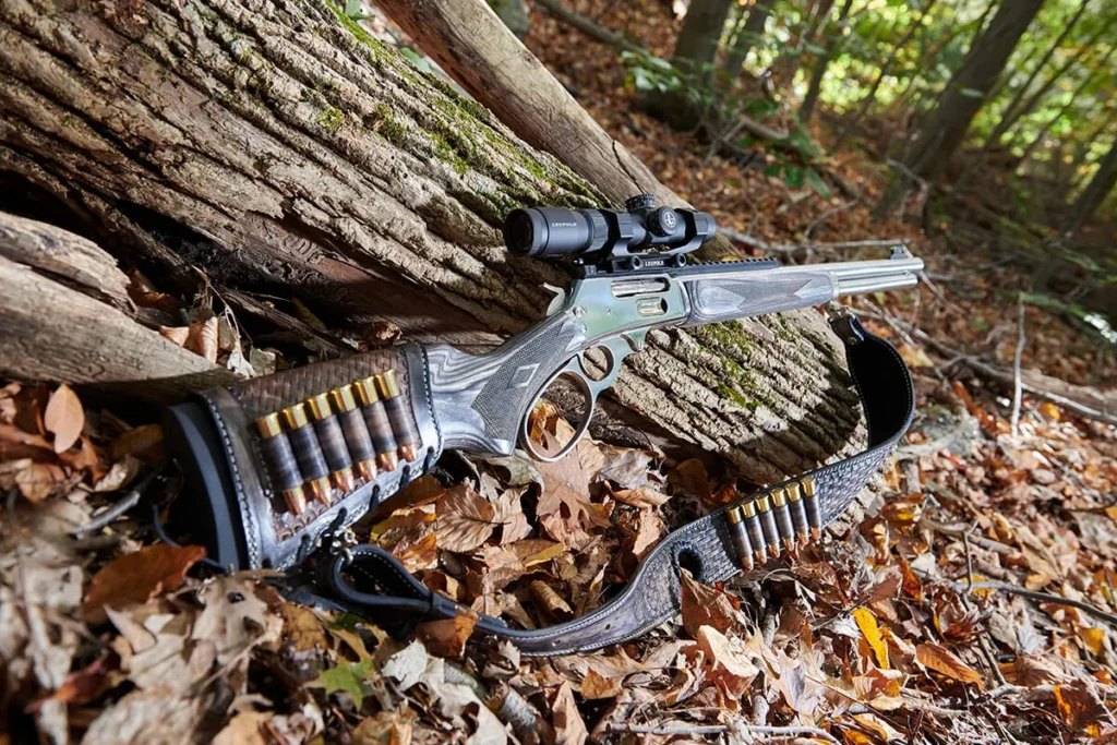 A Marlin 1895 SBL lever-action rifle with a scope, a leather buttstock cartridge holder full of ammunition, and a sling, resting on a log and fallen leaves in a forest setting.