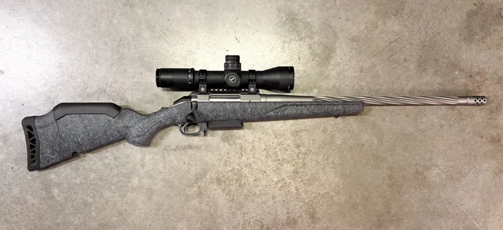 A Ruger American Generation 2 rifle with a gray textured stock, stainless steel fluted barrel, and a large black scope mounted on top, lying on a concrete surface.