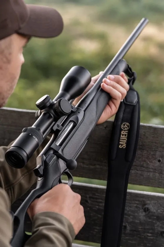 A hunter, viewed from behind the shoulder, holding a Sauer 505 Synchro XT rifle with a large scope attached, showcasing the rifle's design and features.