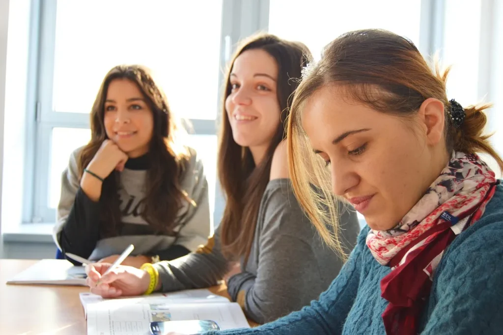 Three young women in a bright classroom setting, one focusing on a book while the others listen or interact, symbolizing learning, discussion, and understanding correct pronunciation.