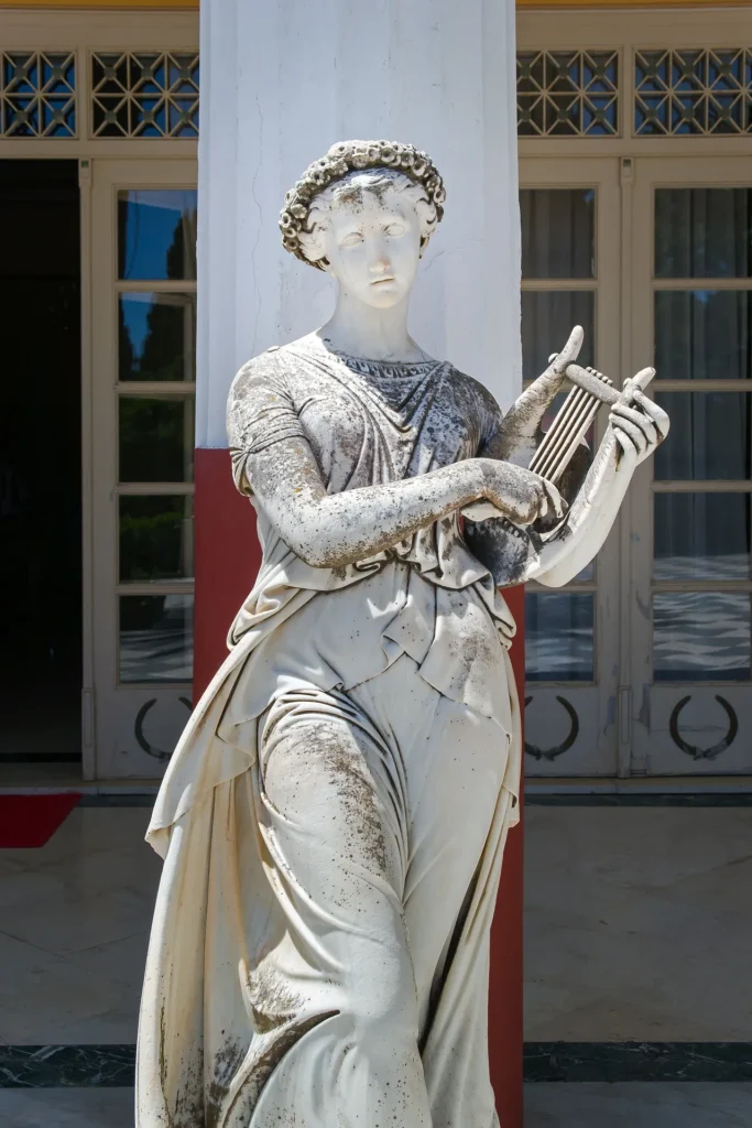 A weathered white marble statue of a classical woman wearing a flowy stola and a floral crown, holding a small lyre against a backdrop of a white pillar and glass doors.