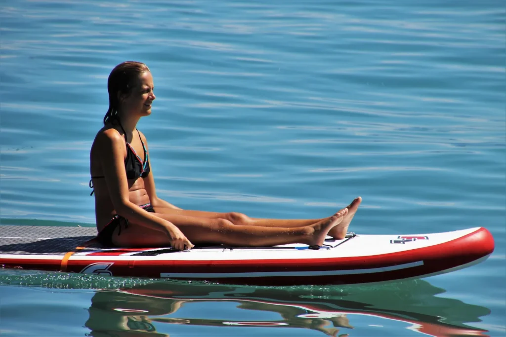 A young woman sitting on a paddleboard in calm blue water, representing modern beauty standards focused on fitness, health, and outdoor lifestyle.