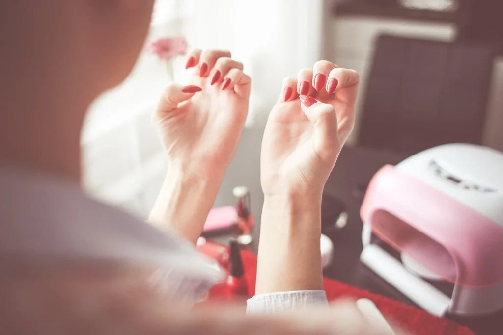 A first-person perspective image showing a person admiring a perfect, high-gloss red gel manicure over their shoulder, with professional nail equipment like a pink and white UV lamp and bottles blurred on the desk below.